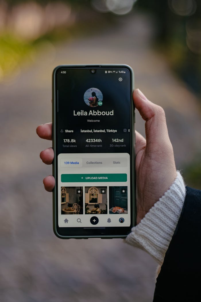 Person holding smartphone displaying user profile, outdoors with bokeh background.